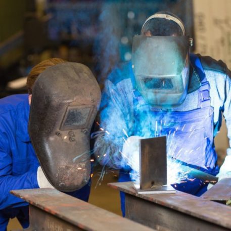 Accompagnement pratique par nos formateurs soudeurs. Formateur et élève avec masques de protection réalisant une soudure sur acier en atelier.